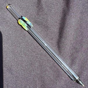 May include: A clear glass pen with a black ink tip. The pen has a decorative butterfly design in yellow, blue, and black. The pen is lying on a dark gray surface.