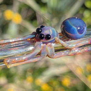 Può includere: Un ragno di vetro con colori iridescenti viola e arancio e occhi neri. Il ragno è seduto su un'asta di vetro trasparente.