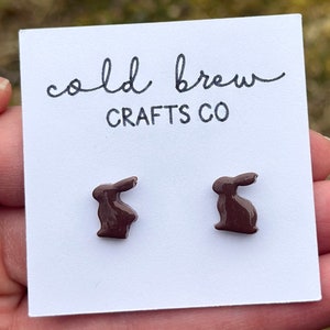 May include: A pair of brown bunny rabbit stud earrings on a white card with the text "cold brew crafts co" printed on it.