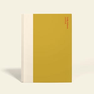 Op de afbeelding: Een mosterdgeel notitieboek met een crèmekleurige rug en een rode verticale streep aan de rechterkant met de tekst "PERSONALISEERBAAR". De notitieboekomslag heeft de tekst "Emily Johnson notebook" in verticale richting.