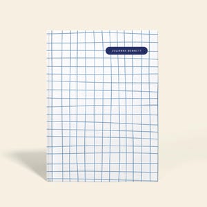 May include: A white notebook with a blue grid pattern. The notebook has a dark blue rectangular label with the name "JULIANNA BENNETT" printed on it. The word "PERSONALIZABLE" is printed vertically on the right side.