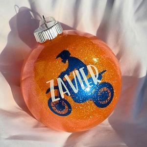 May include: Orange glitter Christmas bauble featuring a blue silhouette of a motocross rider and the name "ZAVIER" in white. The bauble has a silver metal cap.