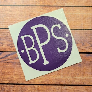 Könnte beinhalten: Ein violetter Kreisaufkleber mit den weißen Buchstaben "BPS" im Inneren. Der Aufkleber befindet sich auf weißem Hintergrund und liegt auf einer Holzoberfläche.