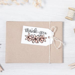 Puede incluir: Una etiqueta de regalo blanca con el texto "thank you" en escritura cursiva negra. La etiqueta est&aacute; decorada con una fila de cinco flores rosas con contornos negros.