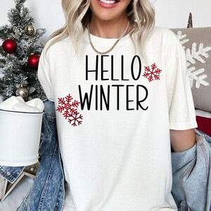 Puede incluir: Camiseta blanca con el texto "Hello Winter" en letras negras y dise&ntilde;os de copos de nieve rojos.