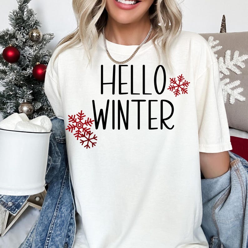 Puede incluir: Camiseta blanca con el texto "Hello Winter" en letras negras y dise&ntilde;os de copos de nieve rojos.