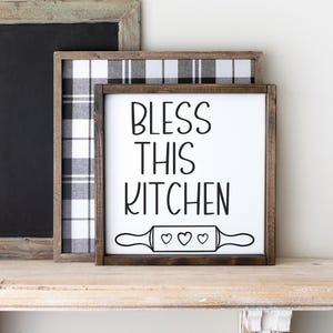 Puede incluir: Un letrero de madera blanco con marco marr&oacute;n que dice "Bless This Kitchen" en letras negras. Debajo del texto hay un rodillo de amasar blanco y negro con tres corazones.