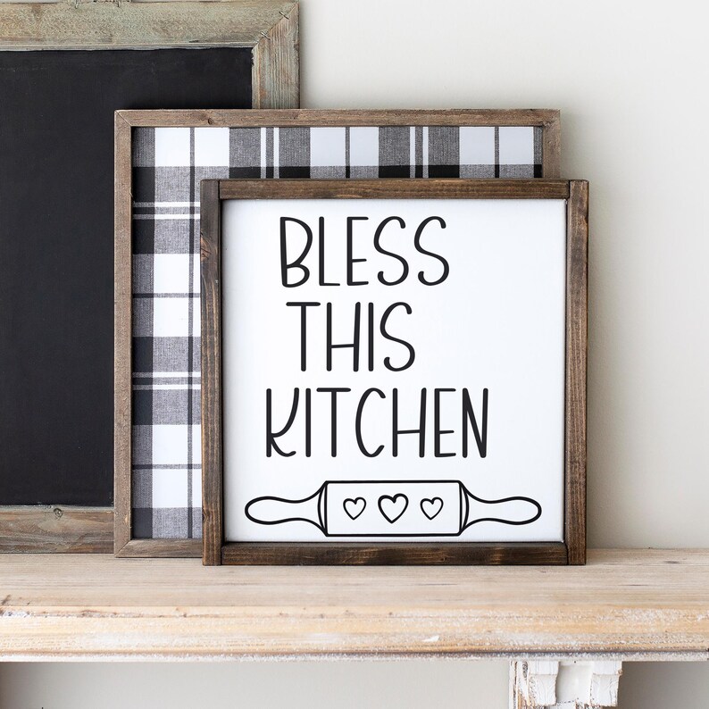 Puede incluir: Un letrero de madera blanco con marco marr&oacute;n que dice "Bless This Kitchen" en letras negras. Debajo del texto hay un rodillo de amasar blanco y negro con tres corazones.