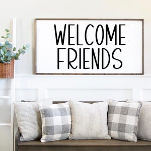 Puede incluir: Un letrero de madera blanco con letras negras que dice "WELCOME FRIENDS". El letrero est&aacute; colgado en una pared blanca sobre un banco de madera con tres almohadas de patrones grises y blancos.