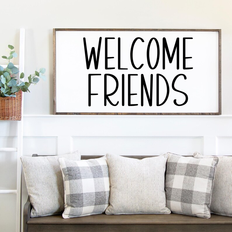 Puede incluir: Un letrero de madera blanco con letras negras que dice "WELCOME FRIENDS". El letrero est&aacute; colgado en una pared blanca sobre un banco de madera con tres almohadas de patrones grises y blancos.