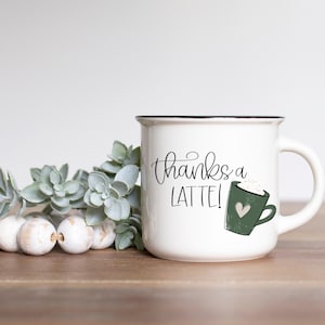 Puede incluir: Taza de cer&aacute;mica blanca con borde negro y una ilustraci&oacute;n de latte verde. La taza tiene el texto "thanks a latte!" en escritura cursiva negra.