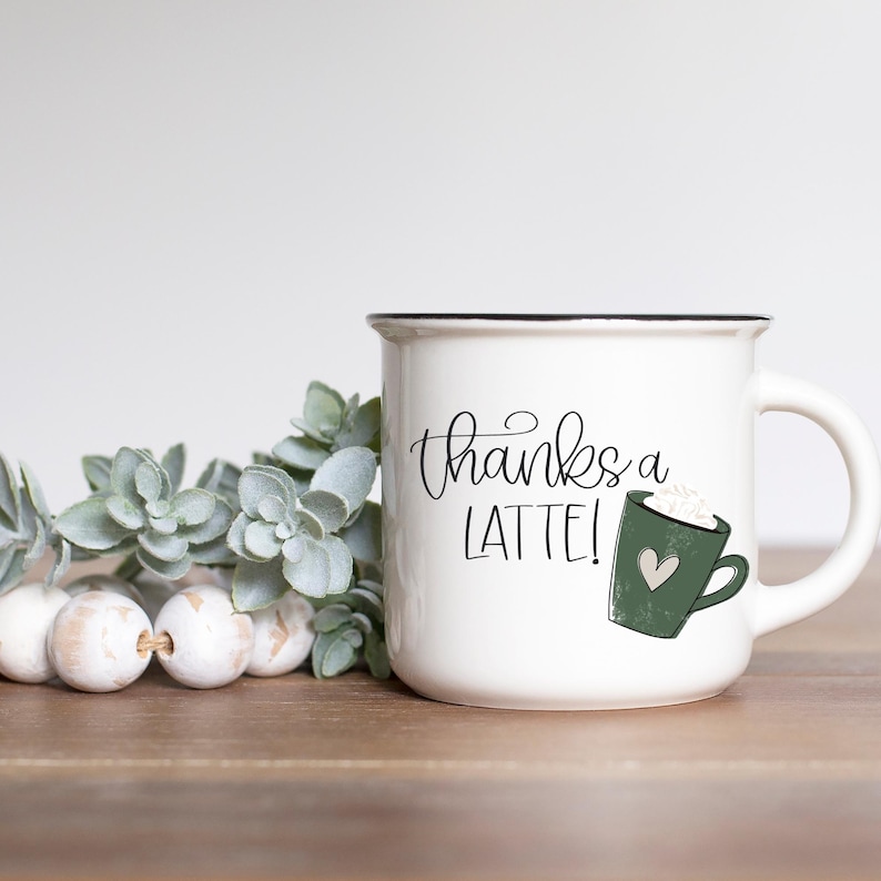 Puede incluir: Taza de cer&aacute;mica blanca con borde negro y una ilustraci&oacute;n de latte verde. La taza tiene el texto "thanks a latte!" en escritura cursiva negra.