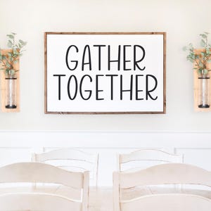 Puede incluir: Un letrero de madera blanco con el texto "GATHER TOGETHER" en letras negras. El letrero est&aacute; colgado en una pared blanca sobre una mesa de comedor con sillas blancas.
