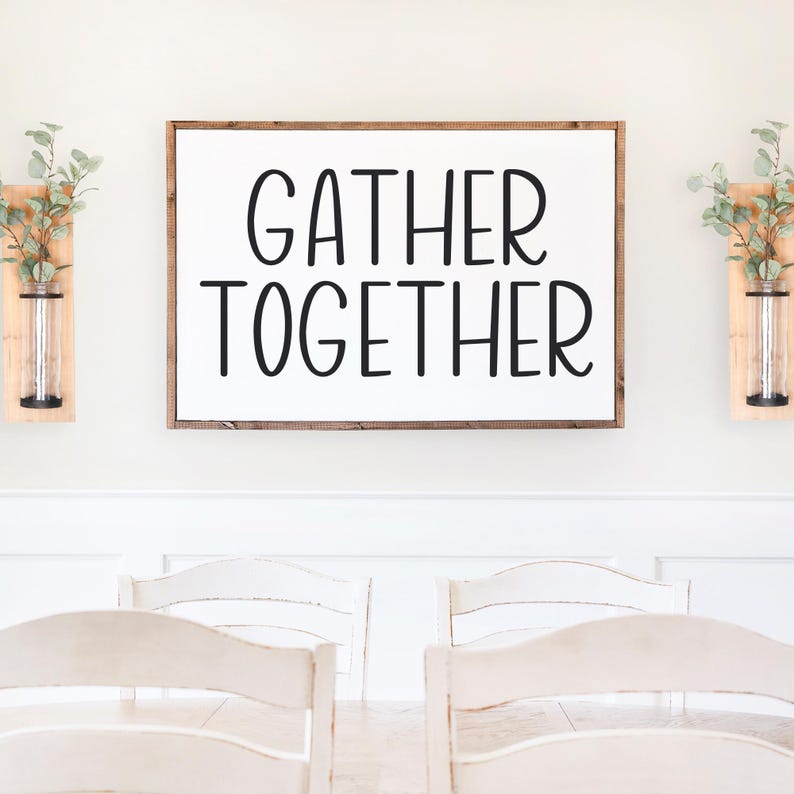 Puede incluir: Un letrero de madera blanco con el texto "GATHER TOGETHER" en letras negras. El letrero est&aacute; colgado en una pared blanca sobre una mesa de comedor con sillas blancas.