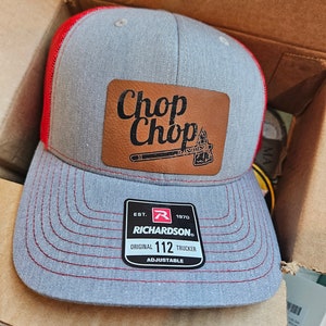 May include: A gray and red trucker hat with a brown leather patch that says "Chop Chop" and a small axe graphic. The hat is adjustable and has the text "Richardson" and "Original 112 Trucker" on the label.
