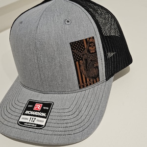 May include: A gray and black trucker hat with a brown patch featuring a skeleton holding a rifle and an American flag. The hat is adjustable and has the text "EST. 1970 RICHARDSON. ORIGINAL 112 TRUCKER ADJUSTABLE" on the label.