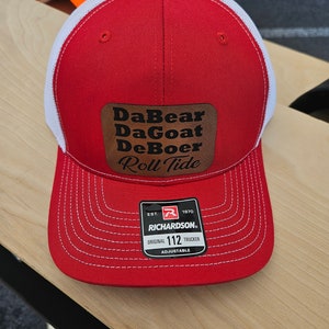 May include: Red and white trucker hat with a brown leather patch that says "DaBear DaGoat DeBoer Roll Tide". The hat is adjustable and has a Richardson 112 logo.