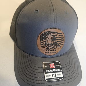May include: Gray trucker hat with a mesh back and a curved brim. The front features a brown leather patch with an eagle and the text "250 YEARS 1776 * 2026". The hat has a Richardson tag.