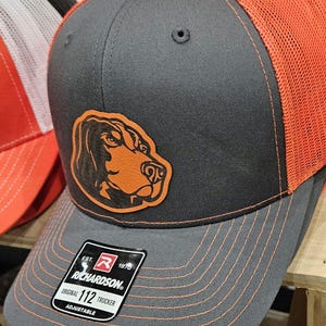 May include: A gray and orange trucker hat with a brown leather patch of a bloodhound's head. The hat has an adjustable strap and a Richardson logo with the text "Est. 1970 Richardson. Original 112 Trucker Adjustable".