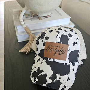 May include: A black and white cow print baseball cap with a brown leather patch that says "Raising Disciples".