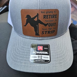 May include: Gray and white trucker hat with a brown leather patch. The patch features a silhouette of a person pole dancing and the text "too young to RETIRE too poor to QUIT too fat to STRIP". The hat has a Richardson 112 tag.