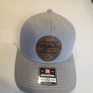 May include: A gray and white trucker hat with a leather patch that reads "America 250" with the years 1776-2026. The hat is a Richardson 112 Original Trucker, adjustable.