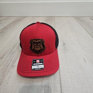 May include: Red and black trucker hat with a brown bulldog logo. The hat has a black mesh back and a red front with a black stitched brim. The logo is a brown bulldog with a black collar and a black chain. The hat has a black label with the text "Richardson. Original 112 Trucker Adjustable" on it.