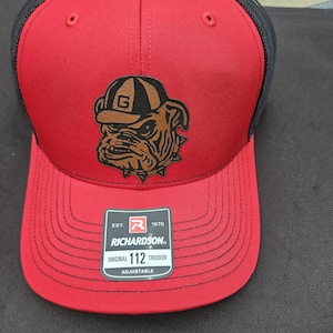 May include: Red and black Richardson 112 trucker hat with a bulldog logo wearing a brown hat. The hat has a curved brim and an adjustable strap. The bulldog logo has a brown and black color scheme. The hat has the text "RICHARDSON ORIGINAL 112 TRUCKER ADJUSTABLE".