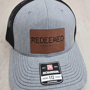 May include: A gray and black trucker hat with a brown leather patch that says "REDEEMED Ephesians 1:7". The hat is adjustable and has a Richardson 112 logo on the back.