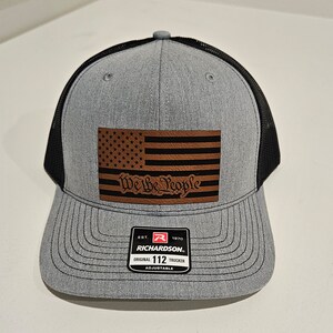 May include: Gray and black trucker hat with a brown leather patch featuring an American flag and the text "We the People". The patch is sewn onto the front of the hat. The hat has a black mesh back and an adjustable strap with a red label that reads "Richardson 112".