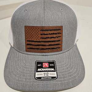 May include: A gray and white trucker hat with a brown leather patch featuring the 2nd Amendment text and an American flag. The patch also includes the text "United States of America". The hat has a black and red logo that says "Richardson" and "Original 112 Trucker Adjustable".