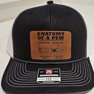 May include: Black and white trucker hat with a brown leather patch that says "Anatomy of a Pew" with a diagram of a bullet and the words "Bang Button", "Freedom Seed", "Metal Holdy Thing", and "Magical Fire Dust". The hat has a white mesh back and a black front with a white stitching detail. The hat has a Richardson 112 label with the text "EST. 1970 RICHARDSON ORIGINAL 112 TRUCKER ADJUSTABLE".