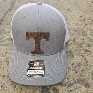 May include: Gray and white trucker hat with a brown leather patch that says "Rocky Top You'll Always Be Home Sweet Home to Me" and "Tennessee". The hat has a white mesh back and a black adjustable strap with the text "Est. 1970 Richardson Original 112 Trucker Adjustable".
