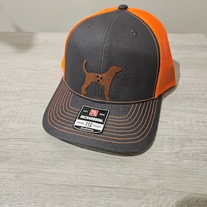 May include: A black and orange trucker hat with a brown embroidered Tennessee Walking Horse silhouette and the text "EST. 1970 RICHARDSON. ORIGINAL 112 TRUCKER ADJUSTABLE" on the front.