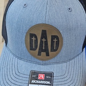May include: A gray and black trucker hat with a brown leather patch that says "DAD" in black letters with two crosses on either side of the "D". The hat has a black mesh back and a label that says "EST. 1970 RICHARDSON. ORIGINAL 112 TRUCKER ADJUSTABLE".