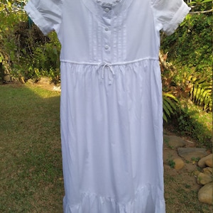 Puede incluir: Un camisón blanco de algodón con mangas cortas, un cuello con ribete de encaje y una falda fruncida. El camisón tiene un cierre de botones en la parte delantera y un lazo en la cintura.