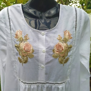 May include: White long-sleeved blouse with a round neckline and button closure. The blouse features delicate lace trim and embroidered floral designs in shades of pink and gold.