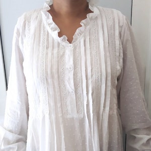 Puede incluir: Camisa blanca de manga larga con cuello con volantes y cierre de botones. La camisa presenta paneles verticales con detalles de encaje y un sutil estampado de lunares. Las mangas son largas y el estilo es suelto y fluido.