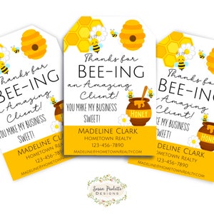 Può includere: Tre etichette regalo gialle e nere a tema ape con il testo "Grazie per essere un cliente fantastico! Rendi il mio business dolce!" e un barattolo di miele con un'ape in cima. Le etichette includono anche il nome "Madeline Clark" e le informazioni di contatto di Hometown Realty.