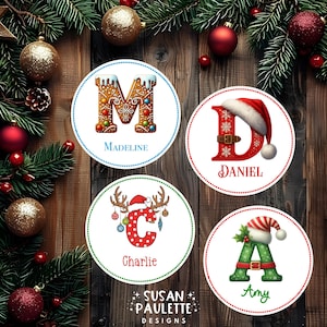 May include: Four circular Christmas-themed labels featuring the letters M, D, C, and A. The M is gingerbread-style, the D is red with a Santa hat, the C has reindeer antlers and ornaments, and the A is green with a striped hat. Names Madeline, Daniel, Charlie, and Amy are printed below each letter.