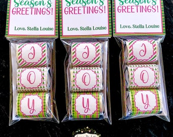 May include: Three clear cellophane bags each containing three individually wrapped chocolate candies. The candies are wrapped in green and pink patterned paper with the letters J, O, and Y printed on them. Each bag has a green and white tag with the text "Season's Greetings! Love, Stella Louise" printed on it.