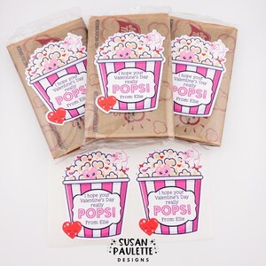 Puede incluir: Bolsas de palomitas de ma&iacute;z para el D&iacute;a de San Valent&iacute;n con pegatinas. Las pegatinas presentan un dise&ntilde;o de cubo de palomitas de ma&iacute;z rosa y blanco con el texto "I hope your Valentine's Day really POPS!" y un coraz&oacute;n. Las bolsas son marrones.