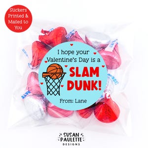 May include: A clear cellophane bag filled with red and silver foil-wrapped chocolate candies. A light blue circular sticker reads "I hope your Valentine's Day is a SLAM DUNK!" with a basketball and net graphic. Text on the sticker says "From: Lane".