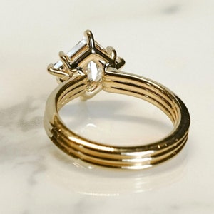 K&ouml;nnte beinhalten: Ein goldener Ring mit einem gro&szlig;en, klaren, rechteckig geschliffenen Mittelstein und kleineren Seitensteinen. Der Ring hat ein dreifaches Banddesign. Die Fassung und das Band sind goldfarben, vor einem wei&szlig;en Hintergrund.