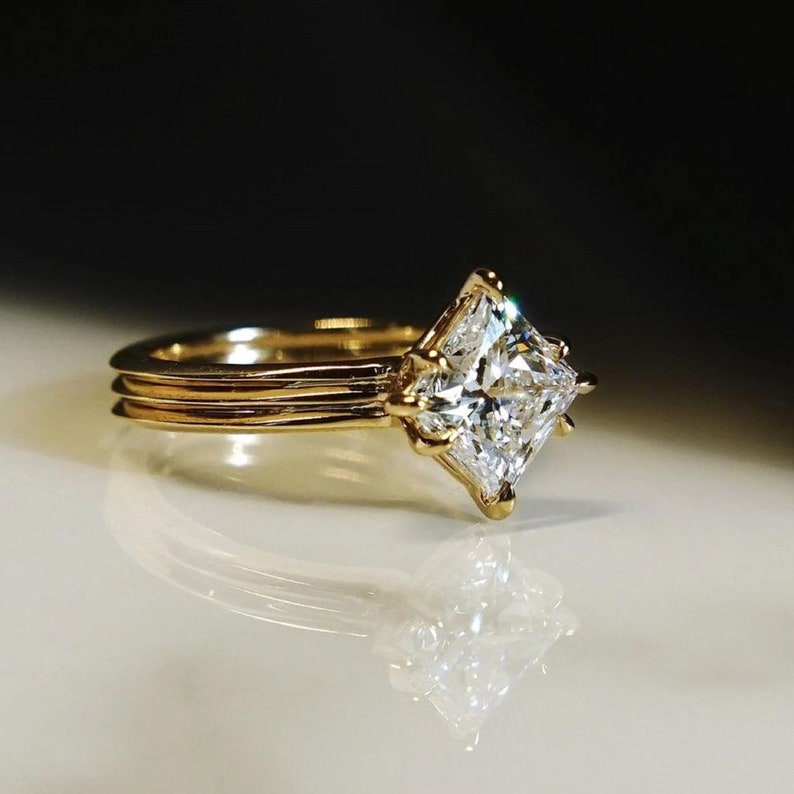 K&ouml;nnte beinhalten: Ein goldener Verlobungsring mit einem gro&szlig;en, quadratisch geschliffenen Diamanten. Der Ring hat ein Doppelband-Design und eine Vier-Zacken-Fassung. Der Diamant funkelt hell vor dunklem Hintergrund.