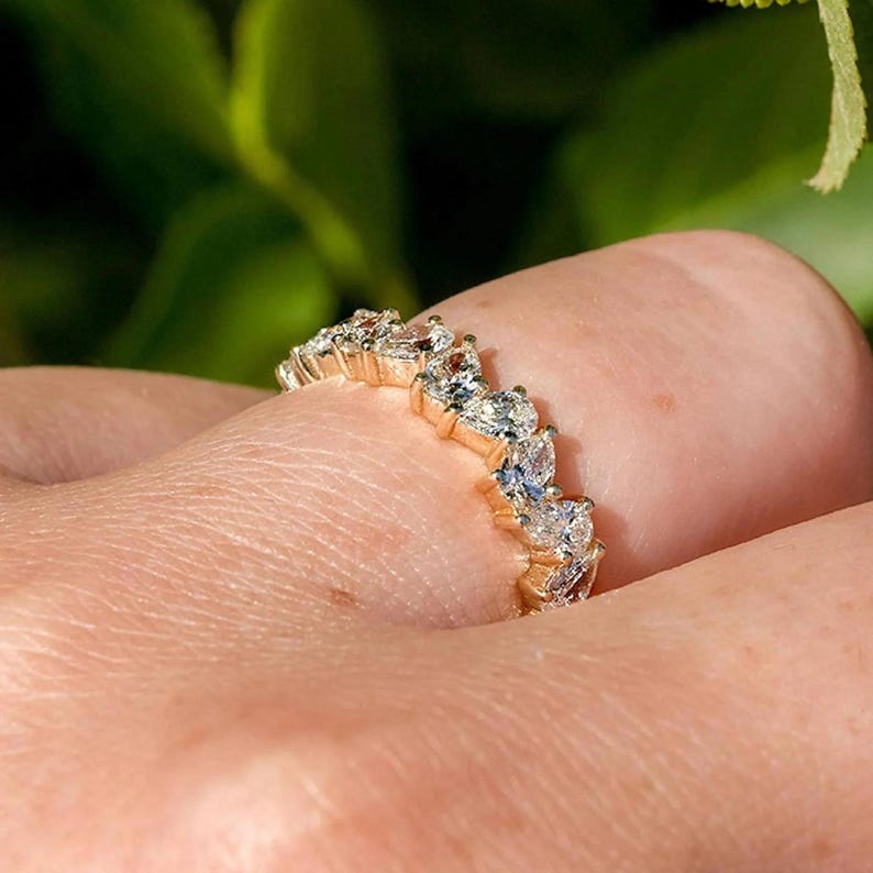 May include: A gold band ring with a pear-shaped diamond setting. The ring is set with multiple diamonds in a continuous line around the band.