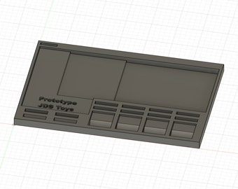 Puede incluir: Una bandeja de almacenamiento de plástico gris con compartimentos y una sección elevada. La bandeja tiene el texto "Prototype JDS Toys" impreso en la parte delantera.