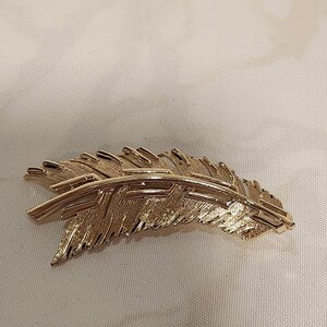 May include: A gold-toned metal brooch shaped like a curved feather. The piece features a central spine with textured, layered barbs extending outward, creating a detailed, realistic representation of a plume with a polished metallic finish.
