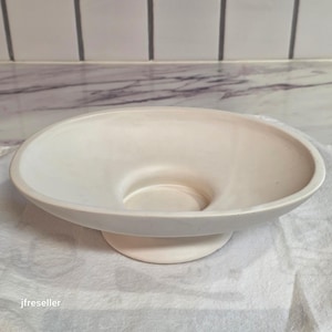 May include: A matte white ceramic bowl with a wide, shallow basin and a circular pedestal base. The bowl features a smooth, uniform finish throughout its interior and exterior surfaces, with a clean, rounded rim and a stable, elevated design.