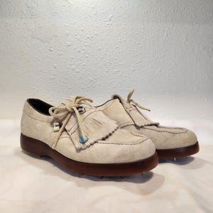May include: A pair of vintage golf shoes in a light beige suede. The shoes have a fringed vamp, brown soles, and metal spikes. Beige laces are tied in a bow. The shoes are a size 8.